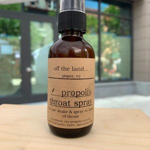 May include: A brown glass bottle of propolis throat spray with a black spray nozzle. The label reads "off the land..." and "propolis throat spray." The ingredients include raw propolis tincture, honey, water, spearmint, and clove.