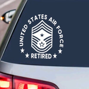 May include: A white vinyl decal sticker with the text "United States Air Force Retired" and a stylized Air Force rank insignia. The sticker is on the back window of a dark blue car.