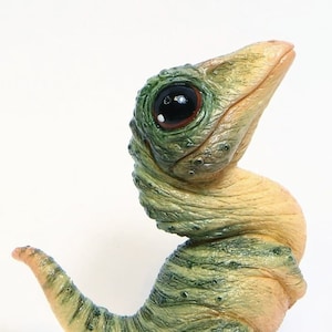 Puede incluir: Una pequeña figura de dinosaurio verde y beige con un gran ojo negro. El dinosaurio está de pie sobre sus patas traseras con la cola enroscada alrededor de su cuerpo.