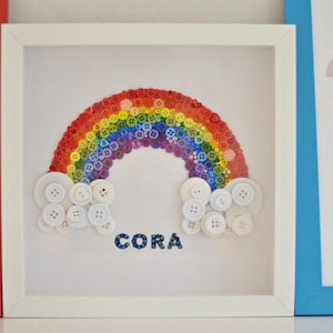 Puede incluir: Una obra de arte enmarcada con un arco&iacute;ris hecho de botones coloridos. Botones blancos forman nubes en la base del arco&iacute;ris, y el nombre "CORA" est&aacute; escrito debajo. La obra de arte est&aacute; en un marco blanco.