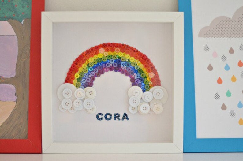 Puede incluir: Una obra de arte enmarcada con un arco&iacute;ris hecho de botones coloridos. Botones blancos forman nubes en la base del arco&iacute;ris, y el nombre "CORA" est&aacute; escrito debajo. La obra de arte est&aacute; en un marco blanco.