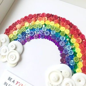 Puede incluir: Una obra de arte enmarcada que presenta un arco&iacute;ris hecho con botones coloridos y gemas brillantes. Nubes blancas en forma de bot&oacute;n est&aacute;n ubicadas en la base del arco&iacute;ris. La obra de arte est&aacute; sobre un fondo blanco.