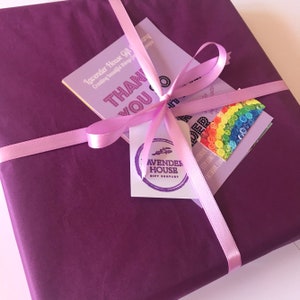 Puede incluir: Un regalo envuelto en papel morado oscuro, atado con una cinta rosa claro. El paquete tiene varias etiquetas, incluyendo una con el texto "THANK YOU" y otra con el logo de Lavender House Gift Company.