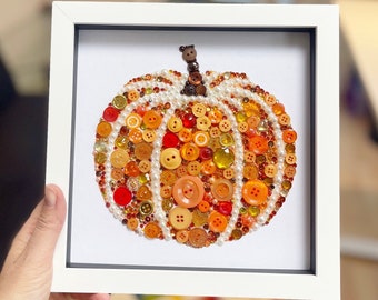 May include: Framed artwork featuring a pumpkin design made from various orange and yellow buttons, pearls, and beads. The pumpkin is set against a white background within a white frame. The stem is crafted from small brown buttons.