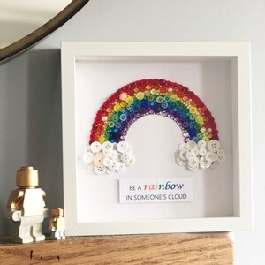 Puede incluir: Una obra de arte enmarcada con un arco&iacute;ris hecho de botones coloridos. Nubes de botones blancos en la base del arco&iacute;ris. El texto "Be a rainbow in someone's cloud" se muestra debajo del arco&iacute;ris.