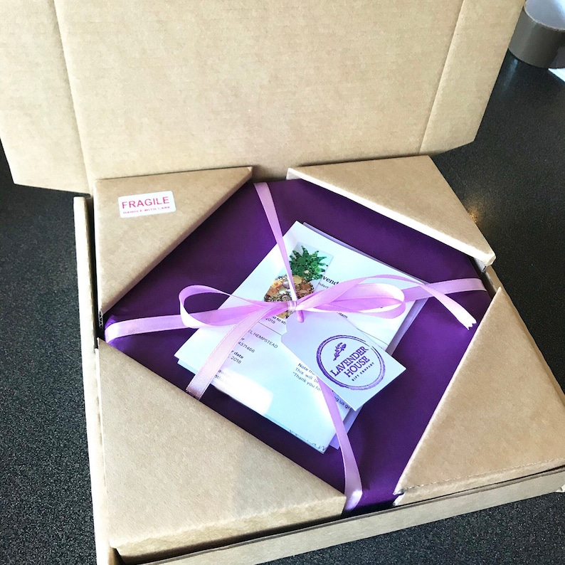 Puede incluir: Un paquete cuadrado envuelto en papel de seda morado y atado con una cinta lavanda, dentro de una caja de cart&oacute;n. El paquete contiene una tarjeta con una ilustraci&oacute;n de pi&ntilde;a y una etiqueta con las palabras "Lavender House Gift Company". La caja tiene una pegatina "Fragile".