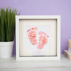 Peut inclure: Une œuvre d'art encadrée avec des empreintes de pieds de bébé faites de boutons roses. Le cadre blanc entoure un passe-partout blanc avec les empreintes de pieds en boutons. Le texte "Hannah 3rd August 2023 3,9 kg" est affiché en dessous.