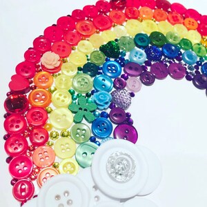 Puede incluir: Un dise&ntilde;o de arco&iacute;ris colorido hecho de varios botones en tonos de rojo, naranja, amarillo, verde, azul y morado. El arco&iacute;ris est&aacute; dispuesto en forma de arco, con botones blancos en la base. Tambi&eacute;n se incluyen cuentas y adornos decorativos.