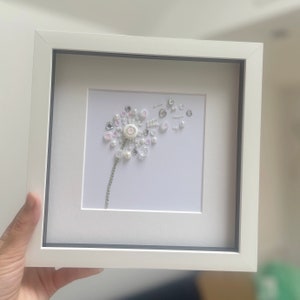 Könnte beinhalten: Gerahmtes Kunstwerk mit einem Pusteblumen-Design. Die Pusteblume besteht aus weißen, transparenten und perlenartigen Perlen und Knöpfen. Der Stiel ist eine silberne Kette. Das Kunstwerk befindet sich vor einem weißen Hintergrund in einem weißen Rahmen.