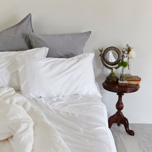 May include: White bedding with a gray pillow and a white pillow on a bed. A small table with a mirror and flowers is next to the bed.