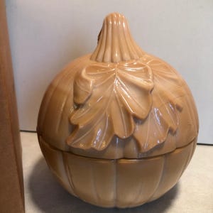 May include: A light peach-coloured glass lidded jar shaped like a pumpkin. The jar features a ribbed exterior surface with a sculpted leaf design on the front and a stem-shaped handle on the lid. The glass has a smooth, glossy finish.