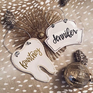 May include: Two white ceramic Christmas ornaments with gold lettering. One ornament is shaped like a tooth and says "Kourtney". The other ornament is shaped like a hat and says "Jennifer".