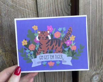May include: A purple card with a cartoon tiger surrounded by flowers. The tiger is orange and black with white stripes. The card has the text "Go get 'em tiger" on a white banner.