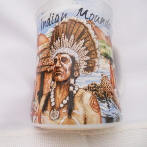 Może przedstawiać: Biały kubek ceramiczny z kolorowym obrazkiem przedstawiającym Indianina w stroju z pi&oacute;rami i naszyjnikiem. Na kubku widnieje napis "Indian Mounds".