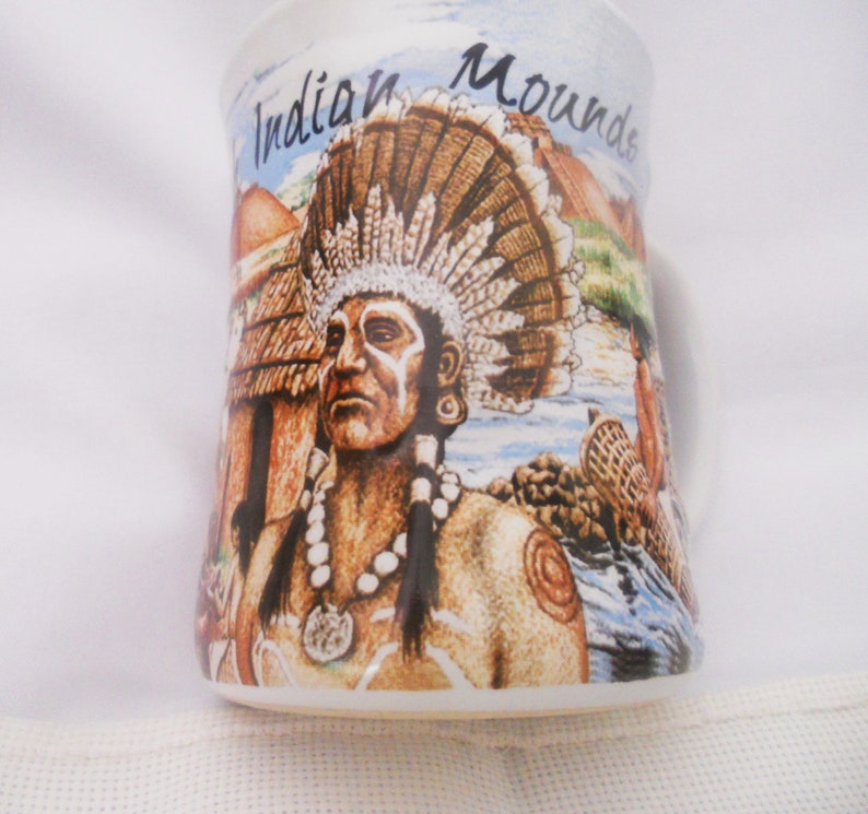 Może przedstawiać: Biały kubek ceramiczny z kolorowym obrazkiem przedstawiającym Indianina w stroju z pi&oacute;rami i naszyjnikiem. Na kubku widnieje napis "Indian Mounds".