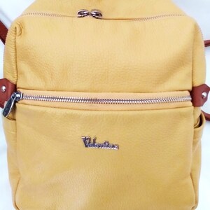 May include: Mustard yellow leather backpack with brown straps and silver zippers. The backpack has a front pocket with a silver zipper and the name "Valentina" in silver lettering. The backpack is a small size.