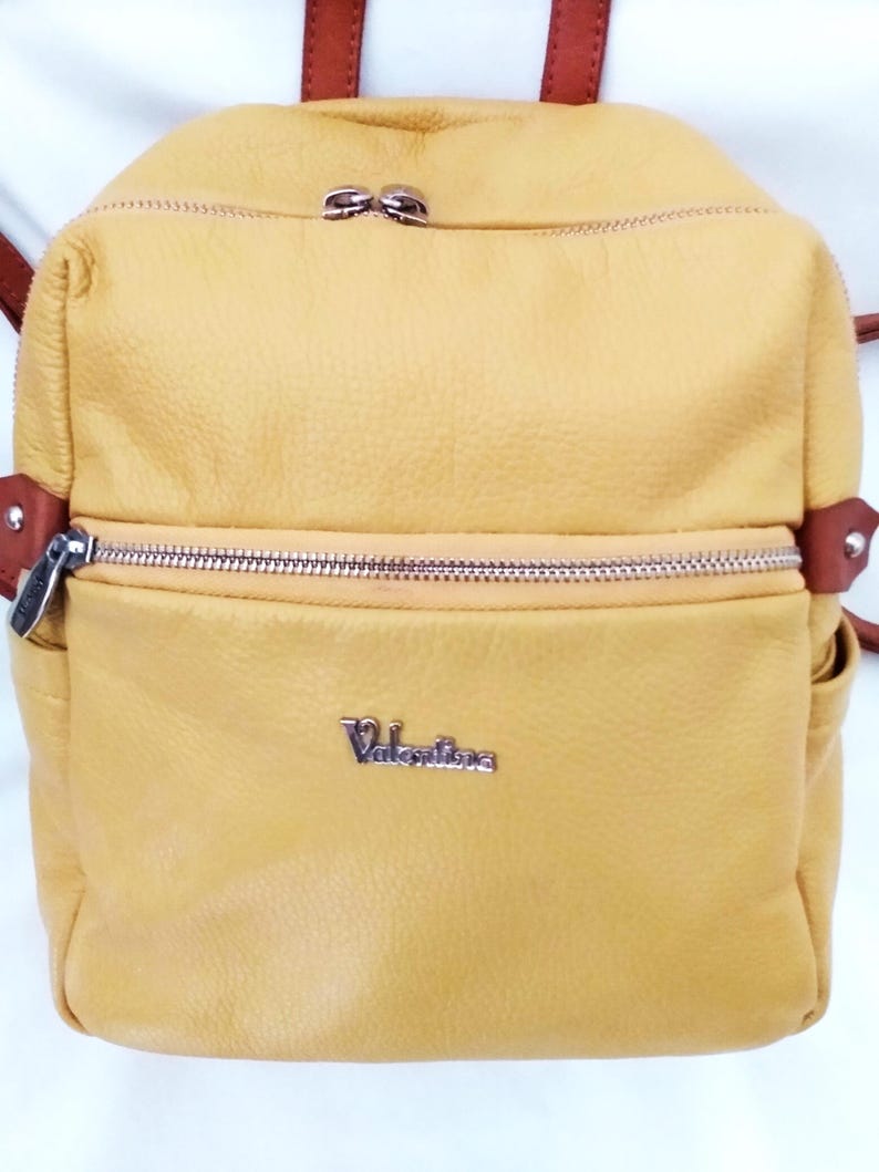May include: Mustard yellow leather backpack with brown straps and silver zippers. The backpack has a front pocket with a silver zipper and the name "Valentina" in silver lettering. The backpack is a small size.