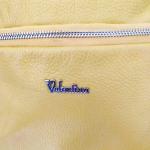 May include: Close-up of a yellow leather bag with a silver zipper. The bag has a textured surface and a silver metal nameplate that reads "Valentina".