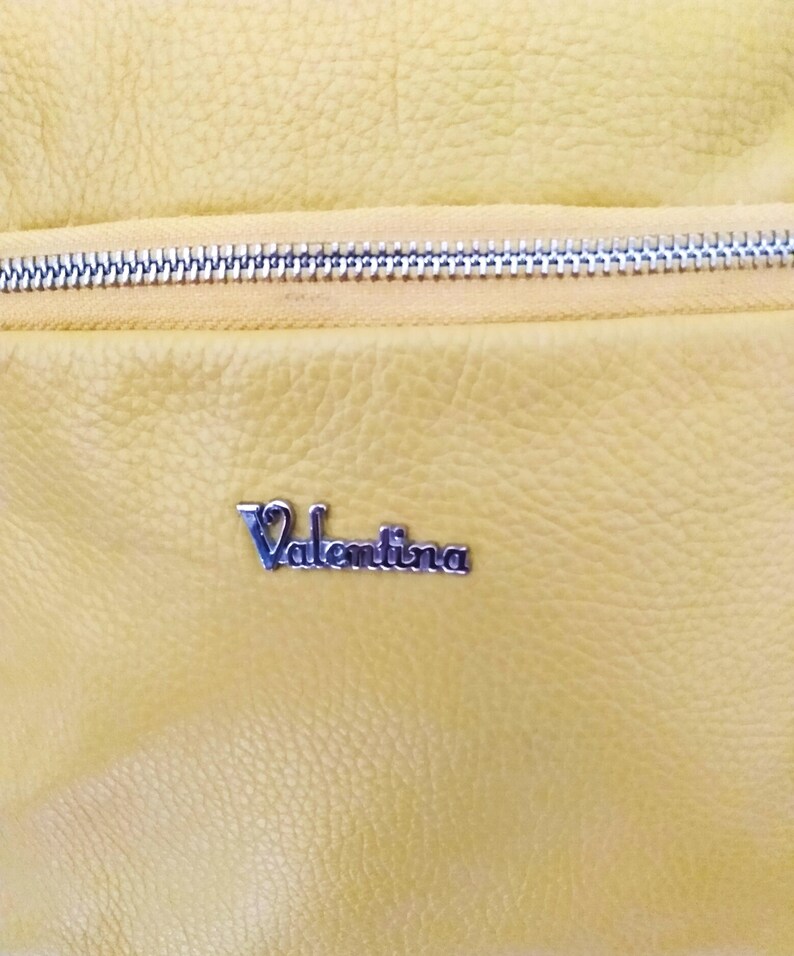 May include: Close-up of a yellow leather bag with a silver zipper. The bag has a textured surface and a silver metal nameplate that reads "Valentina".