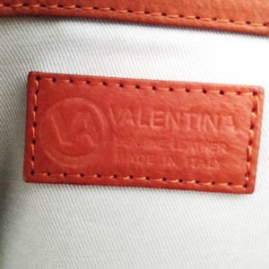 May include: Close-up of a rectangular, orange leather tag with the embossed word "VALENTINA" and a logo. The tag reads "GENUINE LEATHER MADE IN ITALY" and is stitched around the edges. A matching orange leather strap is visible at the top.