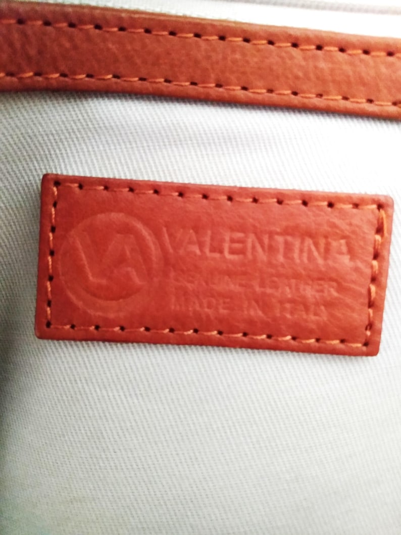 May include: Close-up of a rectangular, orange leather tag with the embossed word "VALENTINA" and a logo. The tag reads "GENUINE LEATHER MADE IN ITALY" and is stitched around the edges. A matching orange leather strap is visible at the top.