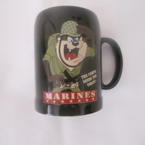 May include: Black ceramic mug featuring a cartoon image of a Tasmanian Devil dressed as a soldier. The mug has the text "MARINES" and "THE CORPS NEEDS YOU TODAY!" printed on it.