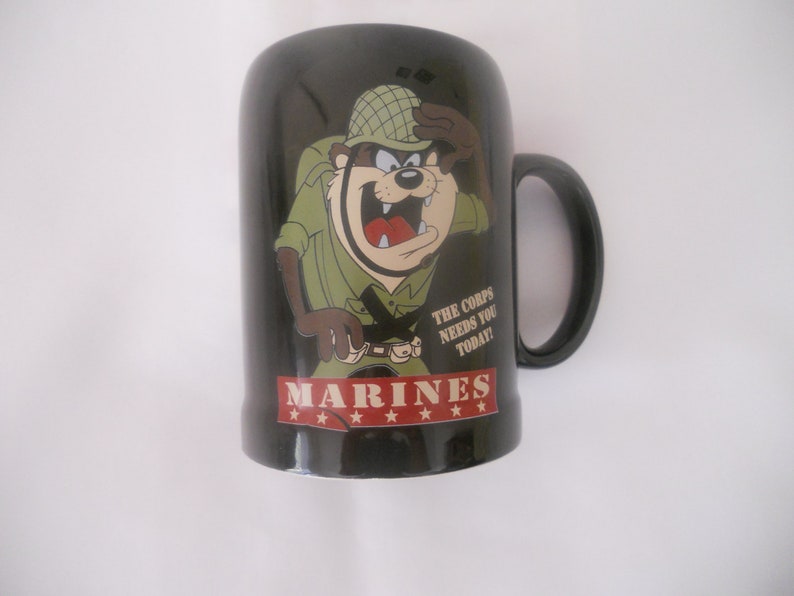 May include: Black ceramic mug featuring a cartoon image of a Tasmanian Devil dressed as a soldier. The mug has the text "MARINES" and "THE CORPS NEEDS YOU TODAY!" printed on it.