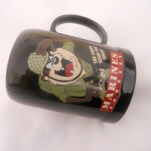 May include: Black ceramic mug featuring a cartoon image of a Tasmanian Devil dressed as a soldier. The mug has the text "The Corps Needs You Today" and "MARINES" with four stars.