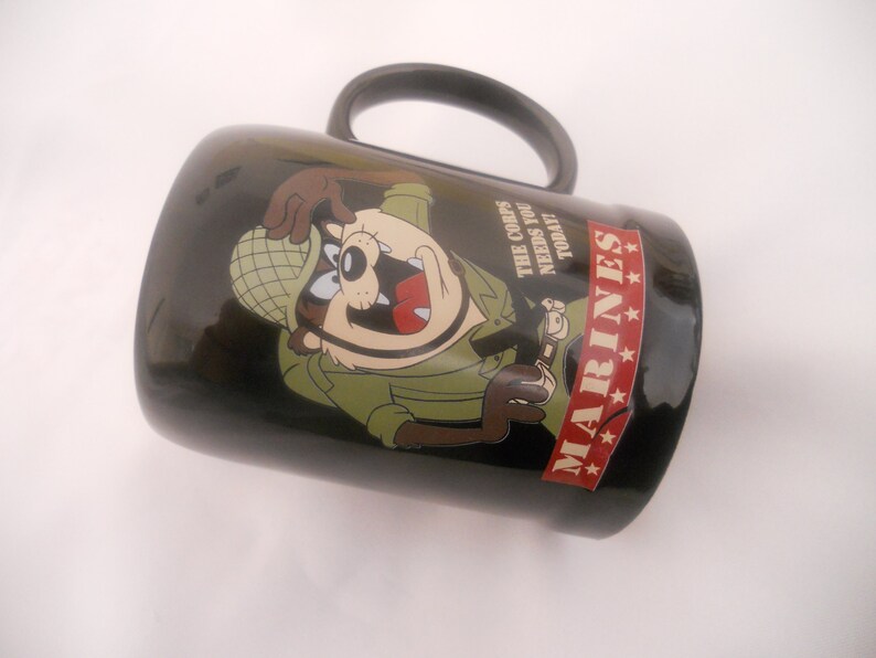 May include: Black ceramic mug featuring a cartoon image of a Tasmanian Devil dressed as a soldier. The mug has the text "The Corps Needs You Today" and "MARINES" with four stars.