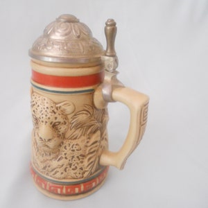 May include: A ceramic beer stein with a detailed image of a leopard and a metal lid with a thumb lift. The stein has a brown and tan base with a red and blue band around the top.