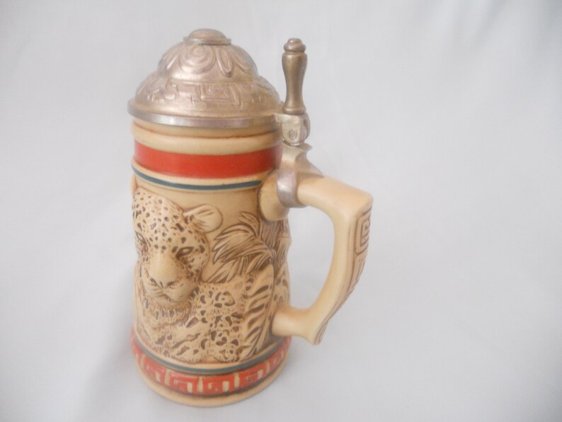 May include: A ceramic beer stein with a detailed image of a leopard and a metal lid with a thumb lift. The stein has a brown and tan base with a red and blue band around the top.