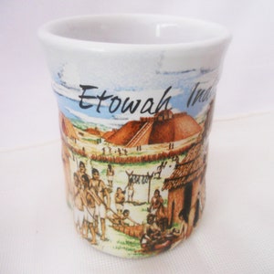 Może przedstawiać: Biały kubek ceramiczny z kolorową ilustracją wioski Indian Etowah. Kubek przedstawia wioskę z domami, ludźmi i dużym kopcem. Na kubku nadrukowany jest tekst "Etowah Indians".