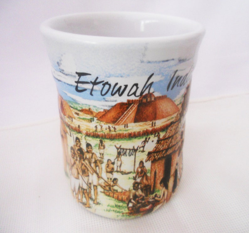 Może przedstawiać: Biały kubek ceramiczny z kolorową ilustracją wioski Indian Etowah. Kubek przedstawia wioskę z domami, ludźmi i dużym kopcem. Na kubku nadrukowany jest tekst "Etowah Indians".