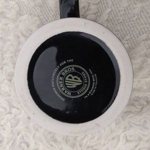 May include: A white ceramic mug with a black base. The base features the Warner Bros. Studio Store logo with the text "Designed Exclusively for the Warner Bros. Studio Store TM & &copy; Warner Bros. 1994 Made in Thailand."