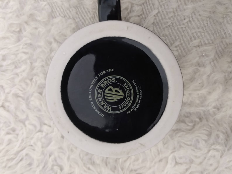May include: A white ceramic mug with a black base. The base features the Warner Bros. Studio Store logo with the text "Designed Exclusively for the Warner Bros. Studio Store TM & &copy; Warner Bros. 1994 Made in Thailand."