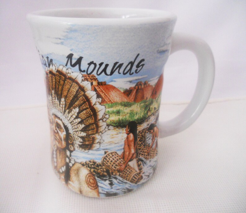 Może przedstawiać: Biały ceramiczny kubek do kawy z kolorowym obrazkiem przedstawiającym rdzennych Amerykan&oacute;w i kopce. Na kubku nadrukowany jest napis "Mounds".