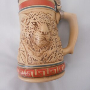 May include: A ceramic beer stein with a detailed relief of a jaguar's face. The stein has a brown and beige color scheme with red and blue accents. The handle is attached to the side of the stein.
