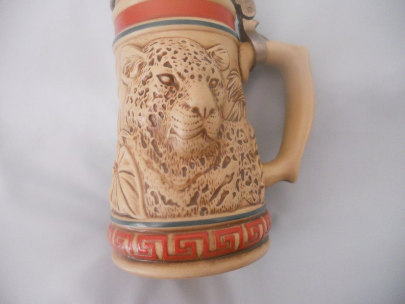 May include: A ceramic beer stein with a detailed relief of a jaguar's face. The stein has a brown and beige color scheme with red and blue accents. The handle is attached to the side of the stein.