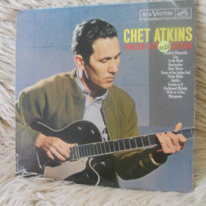 May include: A vintage vinyl record cover featuring Chet Atkins, a renowned guitarist, playing a guitar. The cover is a light blue color with the title "Chet Atkins Finger Style Guitar" in white text. The record includes songs such as "Rhapsody in Blue", "The Mood", "Heartbreakes", and "Gee Whiz".