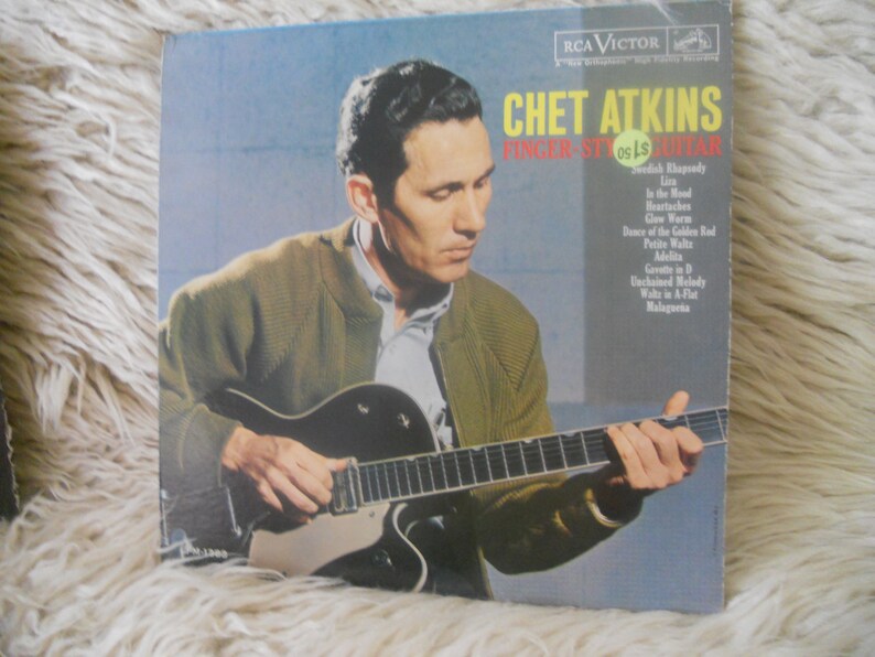 May include: A vintage vinyl record cover featuring Chet Atkins, a renowned guitarist, playing a guitar. The cover is a light blue color with the title "Chet Atkins Finger Style Guitar" in white text. The record includes songs such as "Rhapsody in Blue", "The Mood", "Heartbreakes", and "Gee Whiz".