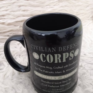 May include: Black ceramic mug with a white graphic design that reads "Civilian Defence Corps". The graphic also includes a star and the text "All Purpose Mug, Crafted with Excellence by The Patriotic Men & Women of Looney Tunes. Standard Issue Item for Civil Defence Members. Our Motto: 'Give 'em ENOUGH and on TIME.'"