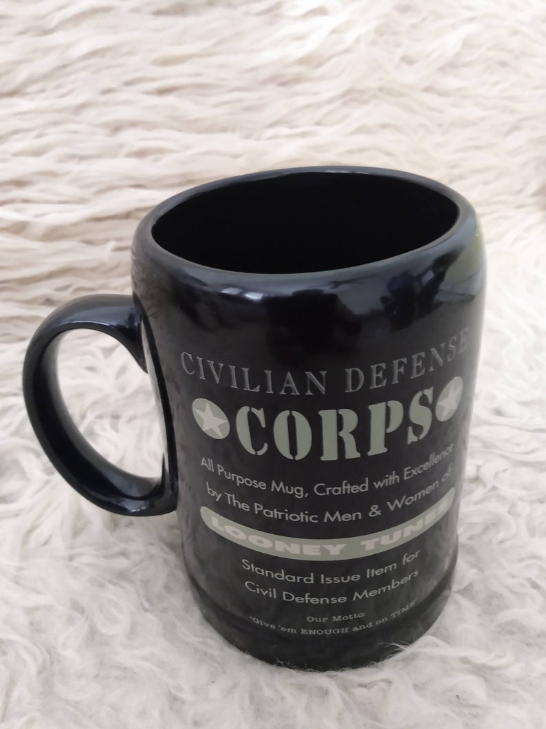 May include: Black ceramic mug with a white graphic design that reads "Civilian Defence Corps". The graphic also includes a star and the text "All Purpose Mug, Crafted with Excellence by The Patriotic Men & Women of Looney Tunes. Standard Issue Item for Civil Defence Members. Our Motto: 'Give 'em ENOUGH and on TIME.'"