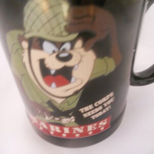 May include: Black ceramic mug featuring a cartoon image of a Tasmanian Devil in a military helmet and camouflage uniform. The text "The Corps Needs You Today" is printed on the mug. The bottom of the mug has the word "MARINES" printed in red with three stars.
