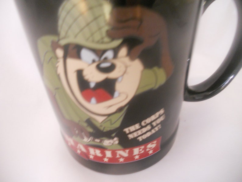 May include: Black ceramic mug featuring a cartoon image of a Tasmanian Devil in a military helmet and camouflage uniform. The text "The Corps Needs You Today" is printed on the mug. The bottom of the mug has the word "MARINES" printed in red with three stars.