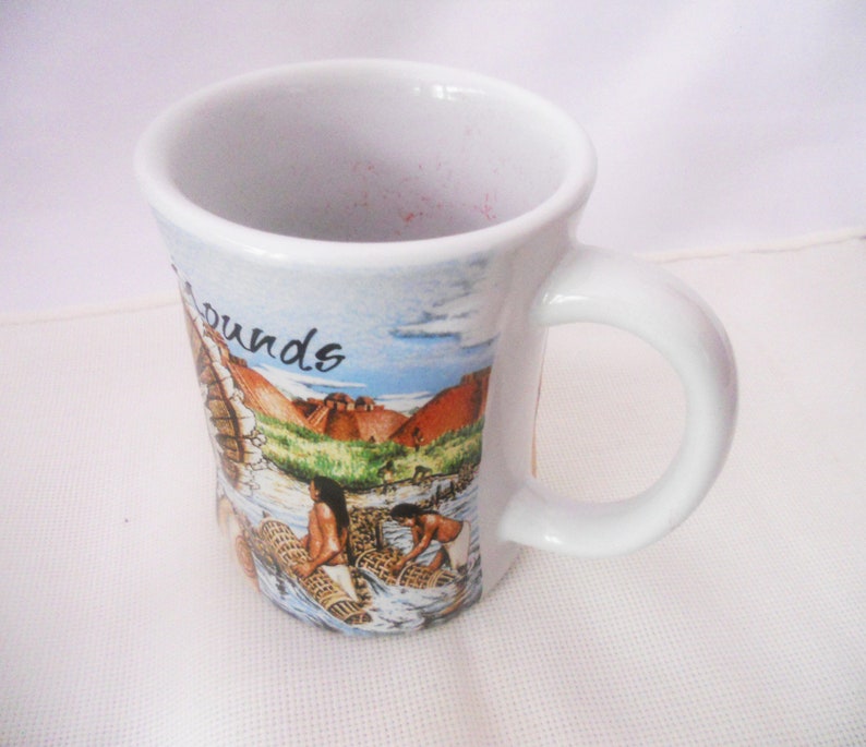 Może przedstawiać: Biały kubek ceramiczny z kolorowym obrazkiem przedstawiającym osoby łowiące ryby sieciami. Na kubku widnieje napis "Mounds".