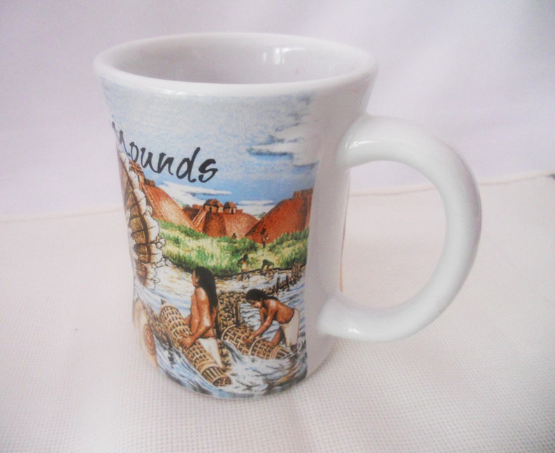 Może przedstawiać: Biały kubek ceramiczny z kolorowym obrazkiem przedstawiającym rdzennych Amerykan&oacute;w łowiących ryby siecią. Na kubku widnieje napis "Mounds".