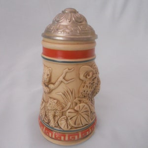 May include: A ceramic beer stein with a silver lid and a relief design of leopards in a jungle setting. The stein has a red and blue band around the middle.