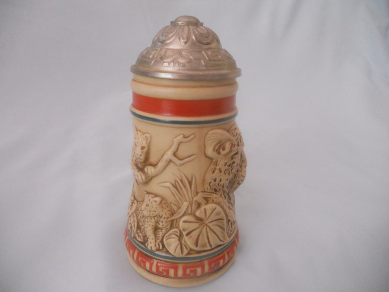May include: A ceramic beer stein with a silver lid and a relief design of leopards in a jungle setting. The stein has a red and blue band around the middle.