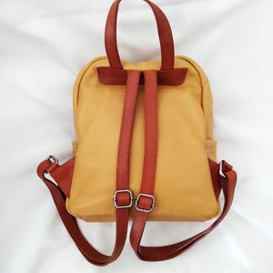 May include: Mustard yellow leather backpack with brown leather straps and silver-colored buckles. The backpack has a rounded shape with a top handle and adjustable shoulder straps. The bag is set against a white background.