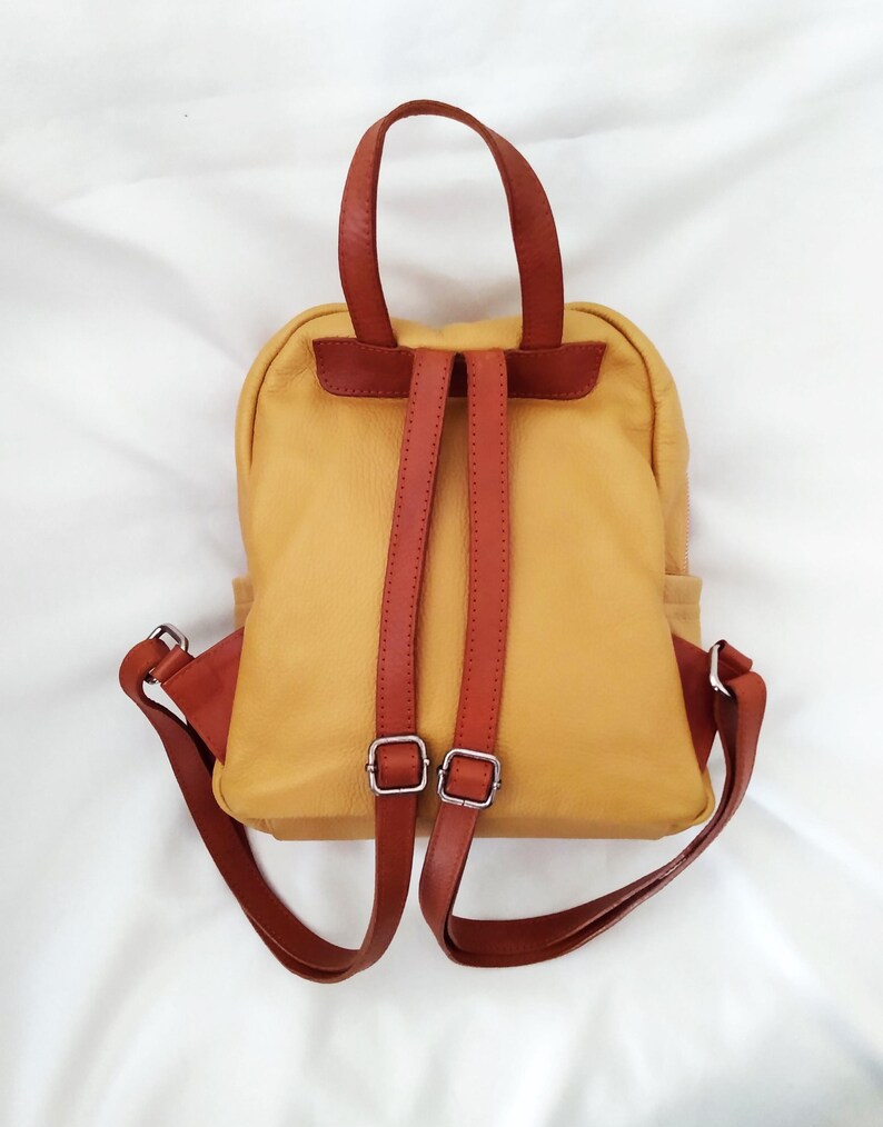 May include: Mustard yellow leather backpack with brown leather straps and silver-colored buckles. The backpack has a rounded shape with a top handle and adjustable shoulder straps. The bag is set against a white background.